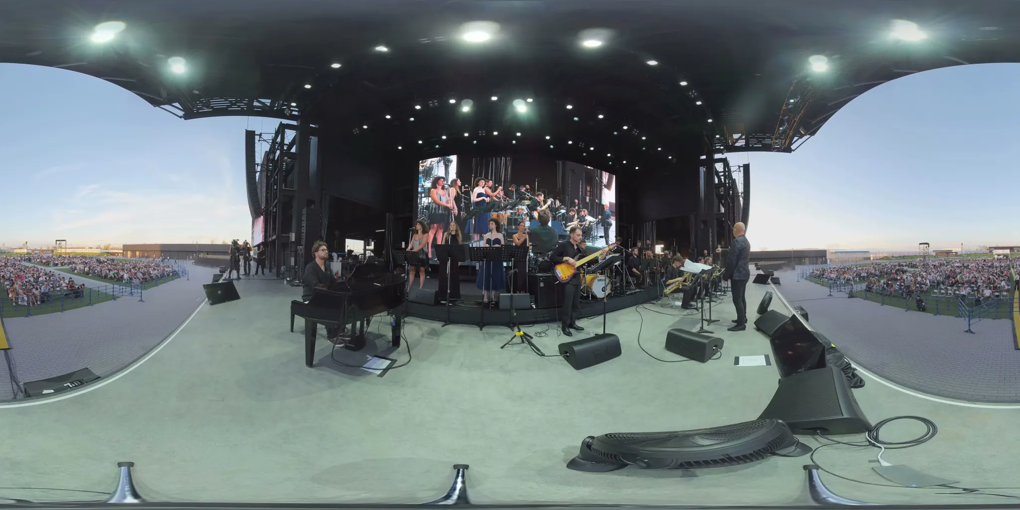 Fotogramma a 360°: un’orchestra jazz su un palco all’aperto, le voci al centro, il pianoforte a sinistra, sassofoni e ottoni a destra, il pubblico alle spalle.