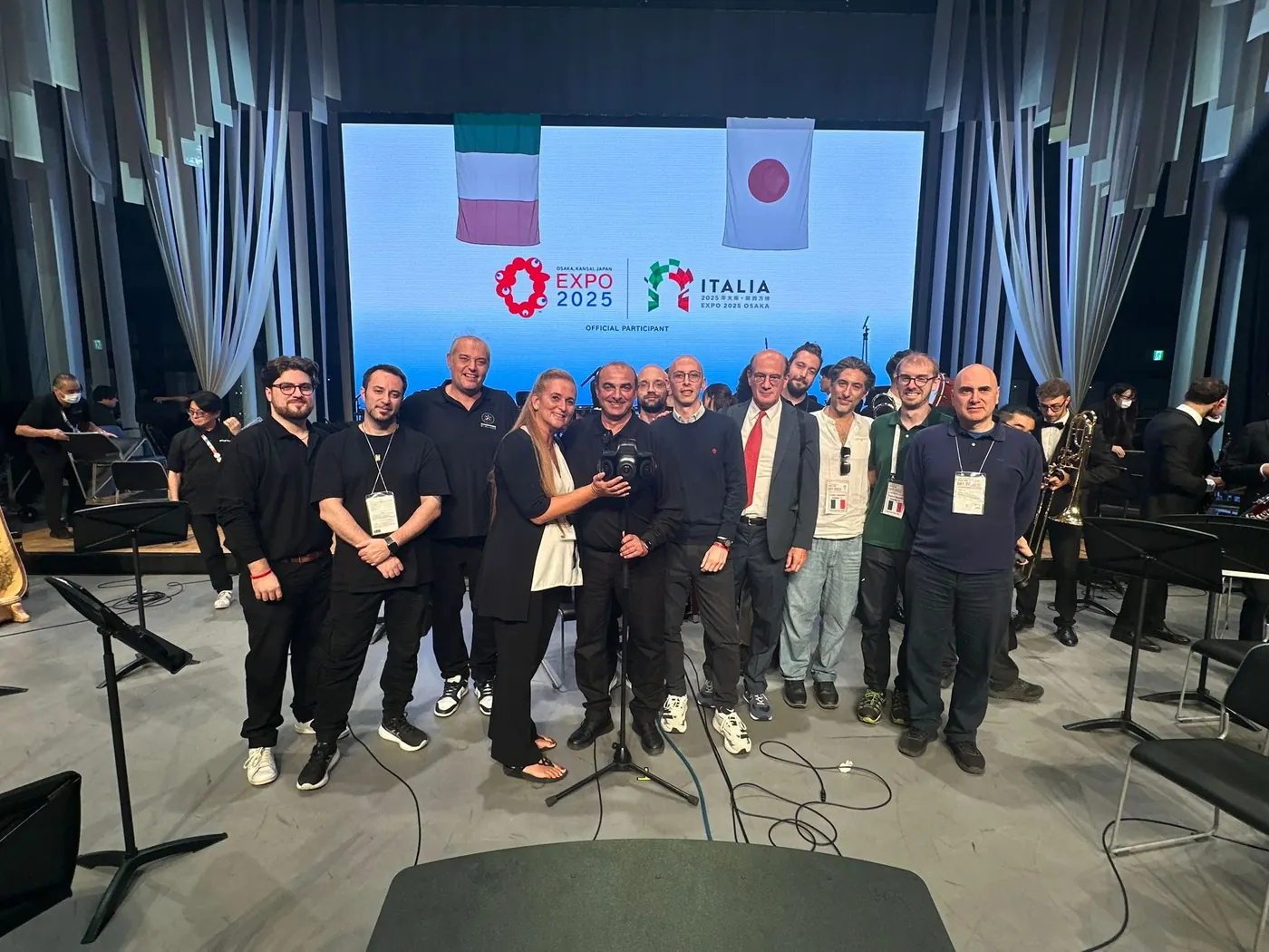 Un gruppo di persone in posa sul palco della National Day Hall, davanti allo schermo con le bandiere italiana e giapponese e i marchi di Expo 2025 e del Padiglione Italia.