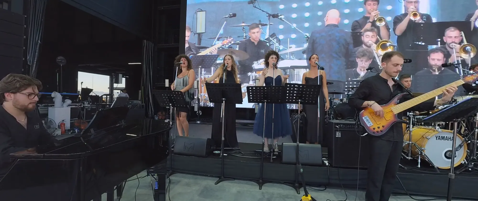 L’Orchestra Nazionale Jazz sul palco dell’Arena Matsuri: il pianista a sinistra, le cantanti ai leggii, il bassista e i fiati, dietro il grande schermo.