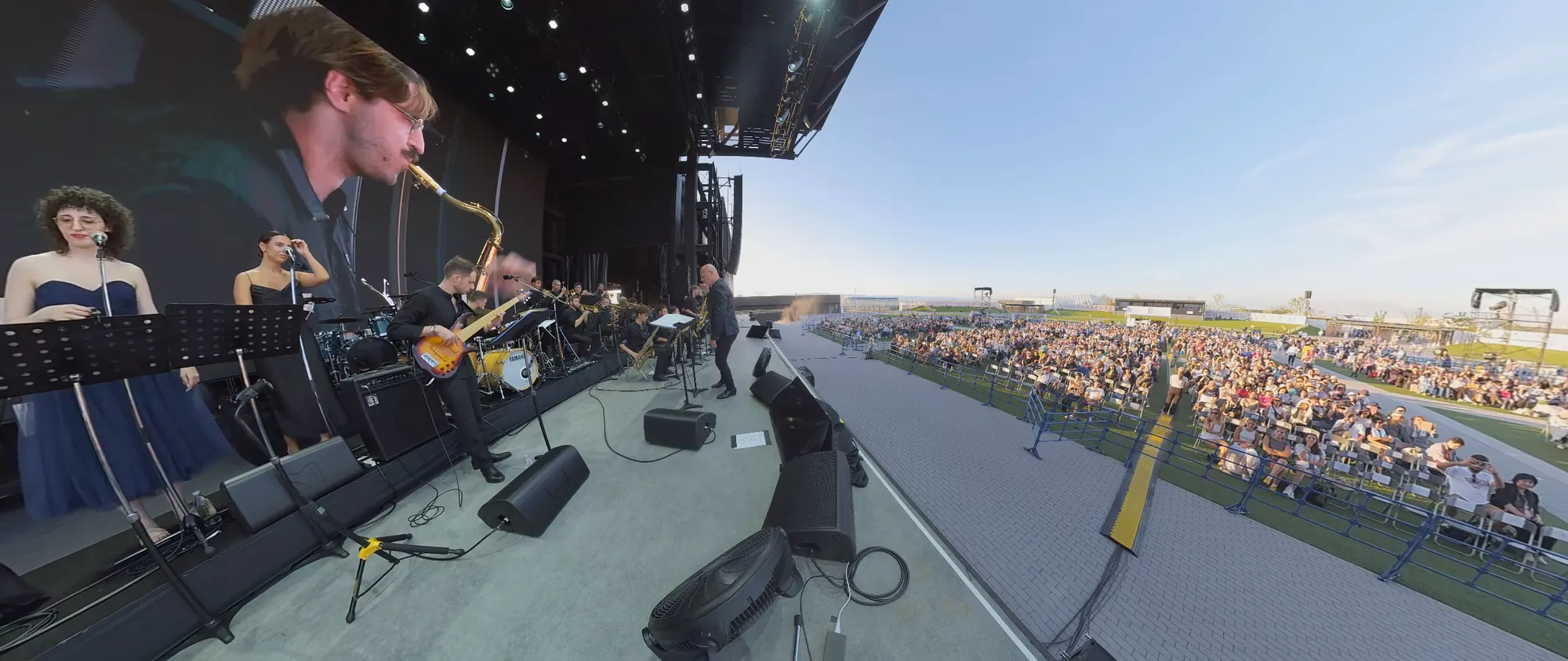 Dal palco dell’ONJ: un sassofonista e le cantanti in primo piano e, oltre il bordo del palco, il pubblico seduto sul prato fino all’orizzonte.