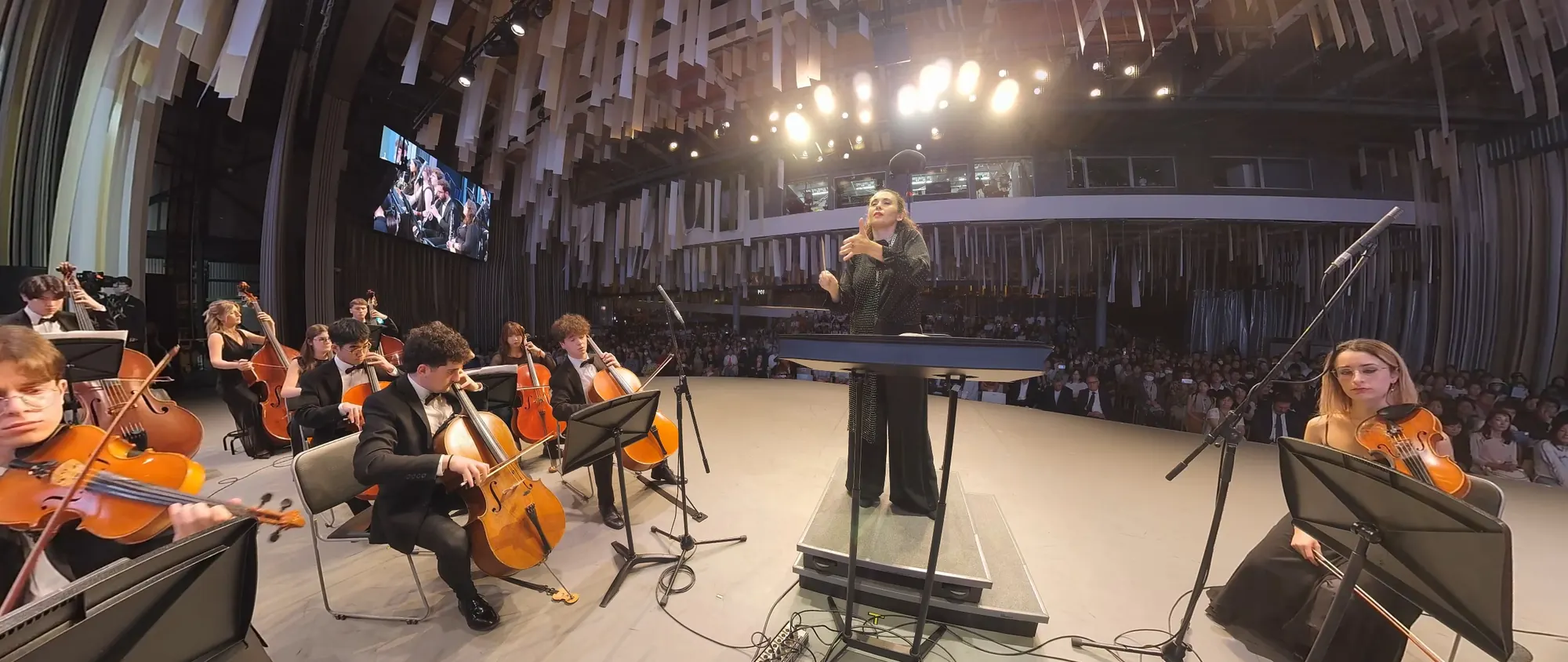 La National Day Hall: la direttrice sul podio, l’orchestra intorno, i lampadari e gli schermi in alto.