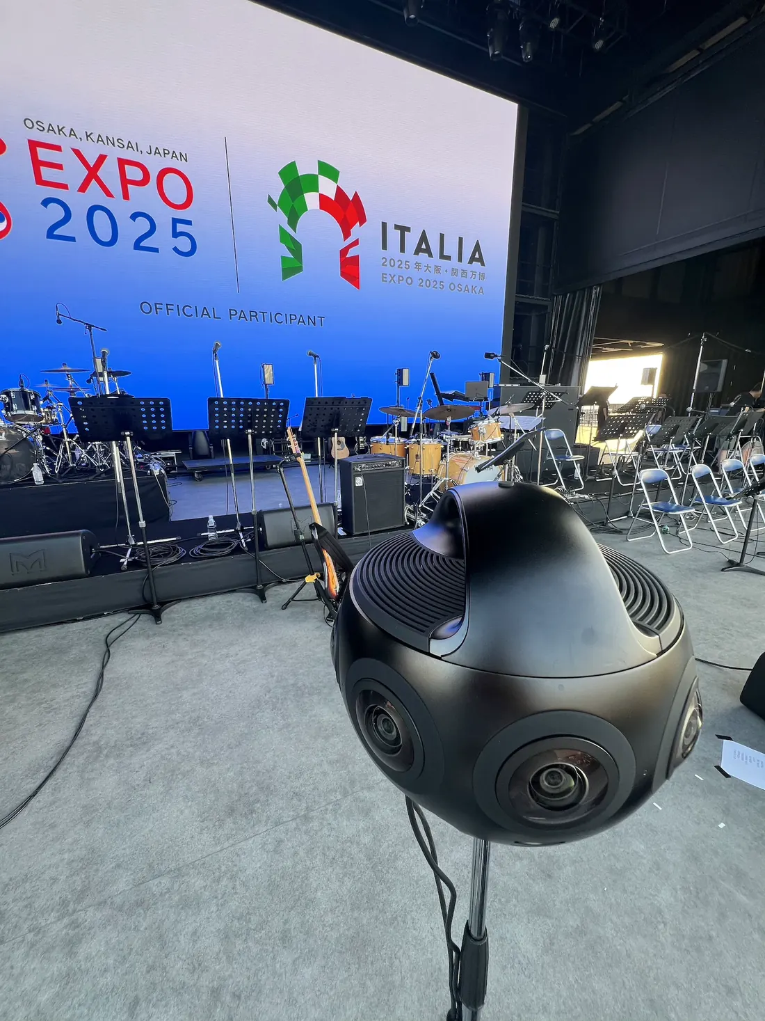 La Insta360 Titan, una sfera nera con otto obiettivi, su un’asta in mezzo al palco; dietro, i leggii vuoti e lo schermo con «Expo 2025 Osaka» e il marchio di Italia.