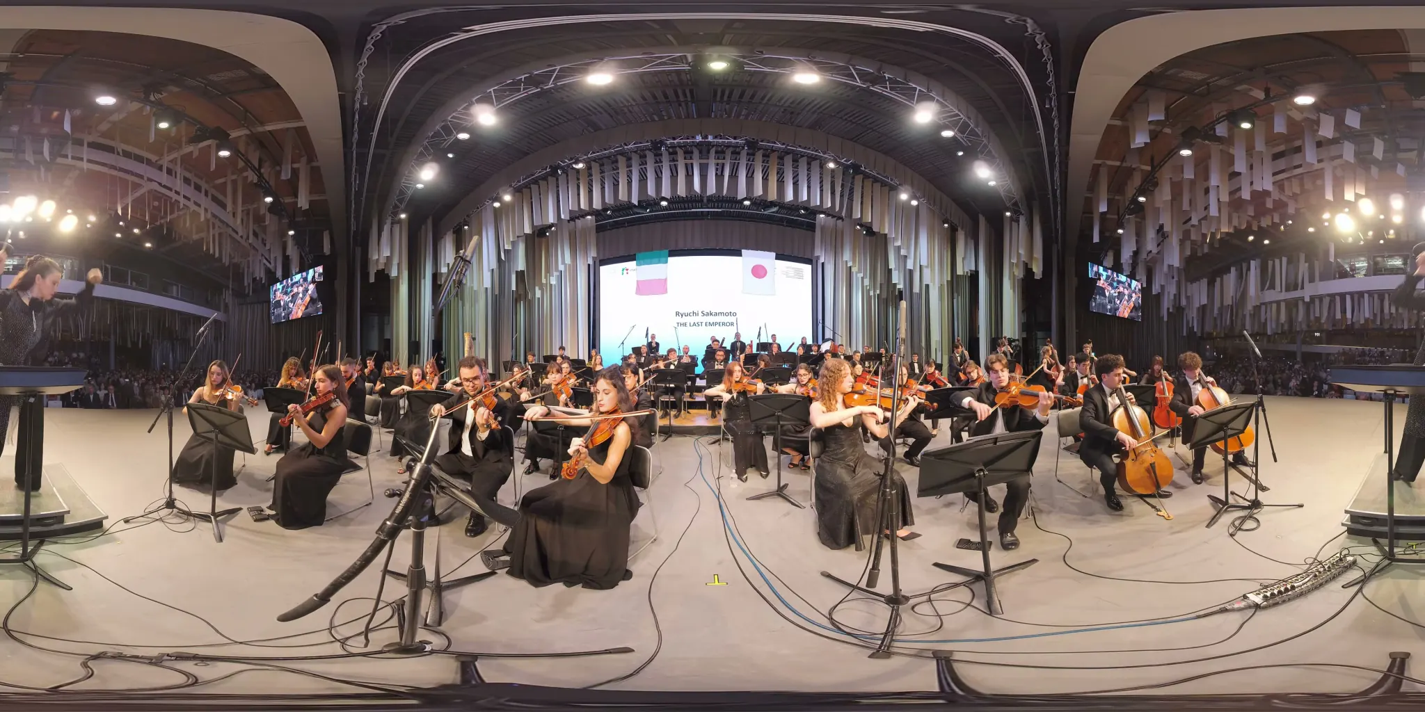 Fotogramma a 360°: un’orchestra sinfonica vista dal podio, i violini a sinistra, i violoncelli a destra; sullo schermo in fondo il titolo «The Last Emperor» e le bandiere italiana e giapponese.